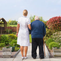 A nurse takes a senior man with walking stick around a garden