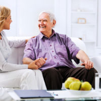 A woman consults with an elderly man sitting on a white sofa